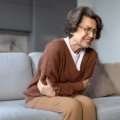 an older woman sits on a sofa clutching her stomach